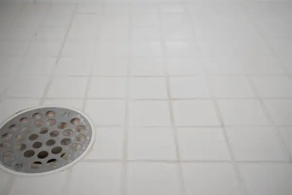 White tiled shower floor with a metal drain cover. The grout lines show signs of wear, indicating a need for tile grout repair.