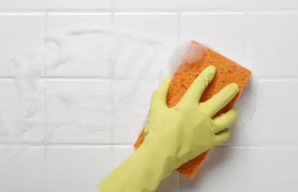 A gloved hand scrubs white bathroom tiles with a soapy orange sponge, showing the best way to clean tiles.
